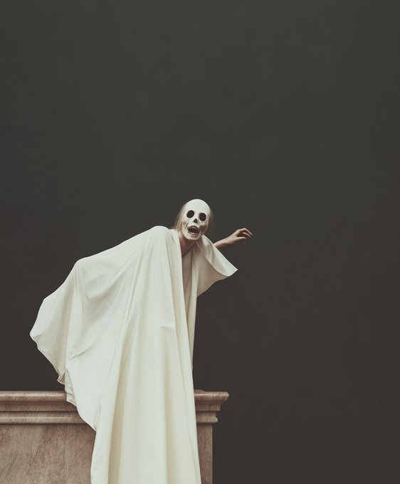 A dramatic, theatrical photograph of a figure in flowing white robes wearing a skull-like mask, posed on a stone ledge against a pitch-black background.