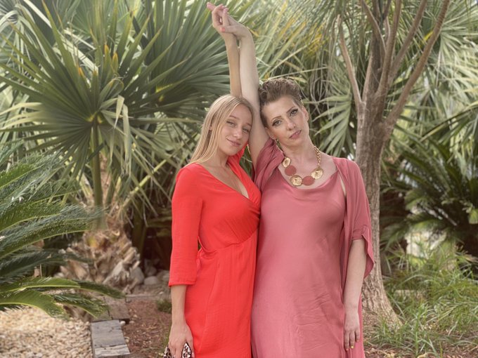 A woman in a coral dress stands closely beside an older woman in pink, arms raised together, posing amid lush palm trees. The image captures a tender moment between what appears to be a mother and daughter, reflecting the poignant post about loss and a complicated relationship.