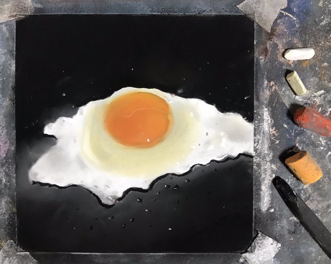 An overhead shot shows a realistic painting of a sunny-side-up egg on a black square surface, accompanied by pastel chalks and a palette knife.