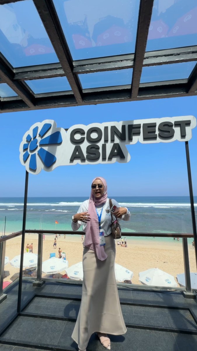 A woman wearing a pink hijab and sunglasses standing under a CoinFest Asia sign at a beachside venue.