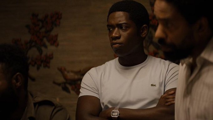 A cinematic still showing a young man in a white t-shirt with crossed arms in a dimly lit room with floral wallpaper, alongside two other men in a serious conversation scene.