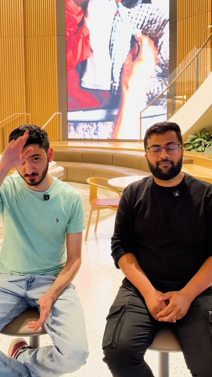 The image features two men sitting next to each other, both wearing black pants and green shirts. They are positioned in front of a large screen that displays an image of a man's face with red hair. The background includes a staircase and a potted plant. The caption for the image reads "Our co-founder @0xsobek teamed up with @yogisomeday to build a zkTLS demo of x402r during the @base <> @privy hackathon thanks for the highlight!".