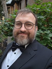 A man with a beard and glasses smiles warmly, dressed in a black tuxedo with a bow tie against a backdrop of greenery.