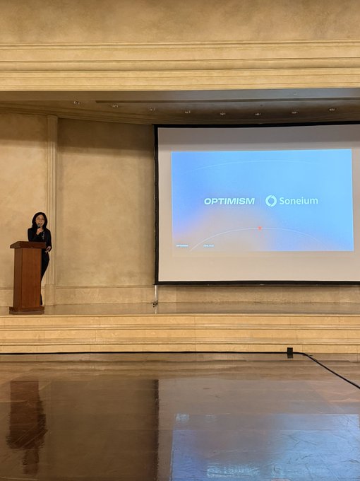 A woman stands at a wooden podium on a stage, presenting alongside a large projection screen displaying the Optimism and Soneium logos. The image captures the early, understated beginning of a significant Web3 collaboration, mirroring the quote's sentiment about pivotal moments often feeling routine at the outset.