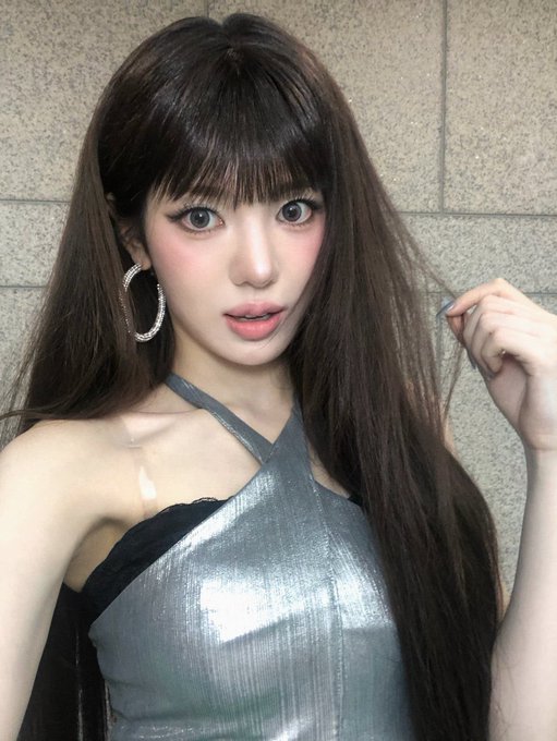 A close-up selfie of a woman with long dark hair and bangs, wearing a silver halter top and sparkly hoop earrings.