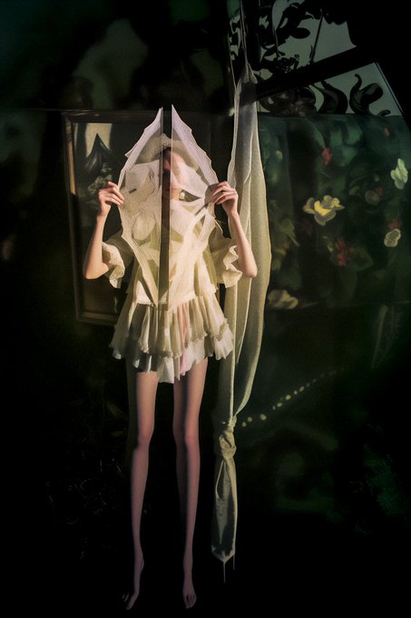 A woman in a flowing cream-colored dress holds sheer fabric draped over her head like a veil in a dimly lit, painterly room with floral artwork. The dreamlike, ethereal scene evokes a sense of quiet emergence or revelation—mirroring the contemplative, threshold-crossing mood of the NFT artwork "Crafting a Door to Dawn."