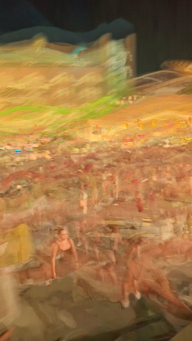 A blurry, motion-smeared photograph showing an indistinct crowd scene with warm yellow and orange tones.