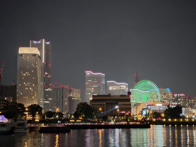 A nighttime cityscape featuring a waterfront view of illuminated high-rise buildings, including a tall white tower, with a green-lit Ferris wheel reflecting on the water.