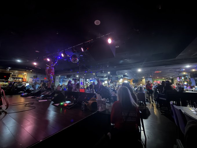 A dimly lit indoor venue, likely a bar or club, with a stage featuring lighting and a disco ball, while a crowd sits and stands watching. A person in the foreground wears a shirt reading "AMANDA K VOGUE".