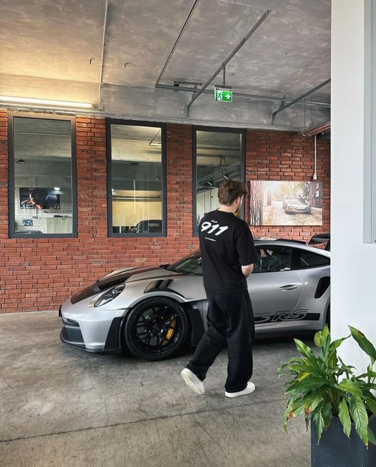 A man wearing a black shirt with '911' on the back walks past a grey Porsche 911 GT2 RS parked in a brick-walled garage.