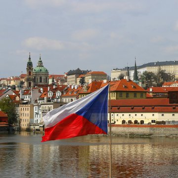 A Czech flag waves in the foreground with Prague's historic skyline, including the Church of St. Nicholas and Prague Castle, visible across the Vltava River. The image visually anchors the news of the Czech Republic's decision to block access to Polymarket, a prediction market platform, by adding it to the country's gambling blacklist.