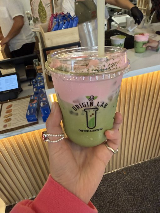 A hand holding a clear plastic cup containing a layered pink and green beverage with chia seeds and a straw, branded with 'ORIGIN LAB COFFEE & MATCHA' at a cafe counter.