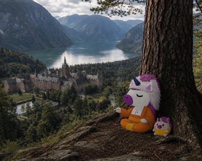Two plush toys—one resembling a unicorn with purple hair and the other a small round character—are posed sitting against a tree trunk overlooking a scenic view of a castle beside a fjord/lake surrounded by mountains.