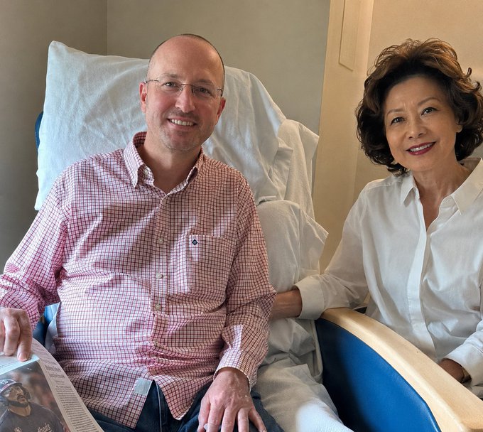 Two people, a man in a red and white checkered shirt and glasses, and a woman in a white blouse, smiling together in what appears to be a hospital room.