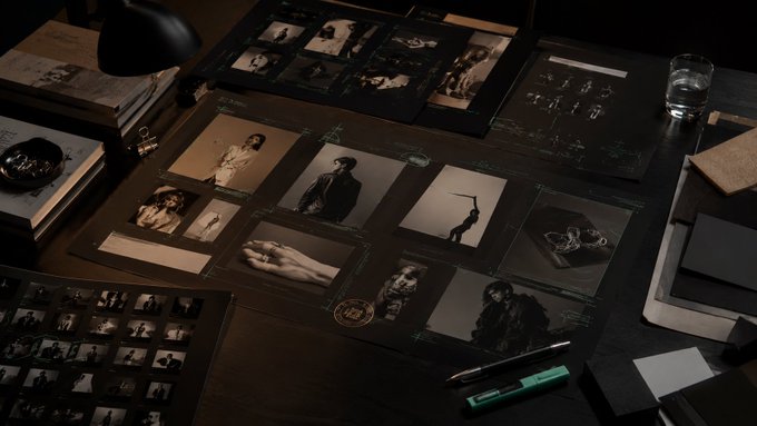A moody, dimly-lit desk displays an array of black-and-white photographs and contact sheets spread across the surface, alongside a lamp, books, a glass of water, and a green marker.