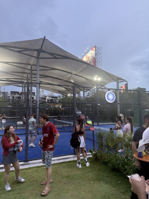 A padel court at dusk with players on the court and spectators standing on the grass nearby, under a white canopy structure.