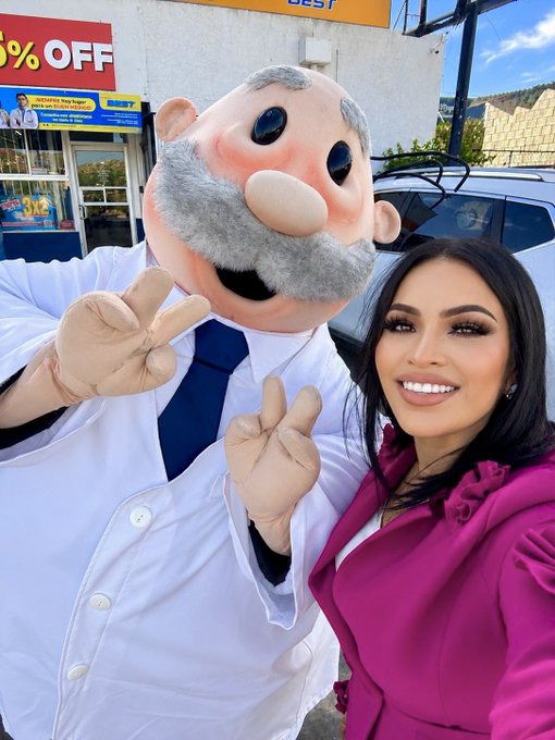 A woman in a pink coat poses for a selfie with a mascot wearing a white coat and blue tie who is making peace signs, taken outside a Best Buy store.