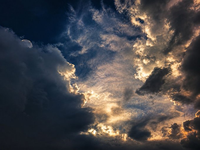 A dramatic photograph of dark, voluminous clouds illuminated by golden sunlight breaking through, set against a deep blue sky.