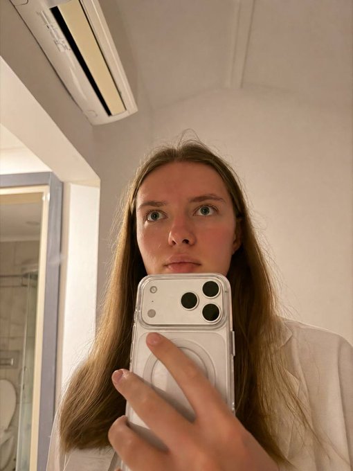 A mirror selfie of a young woman with long blonde hair wearing a white shirt, holding an iPhone in a clear case that displays a MagSafe ring, taken in a room with a wall-mounted air conditioner.