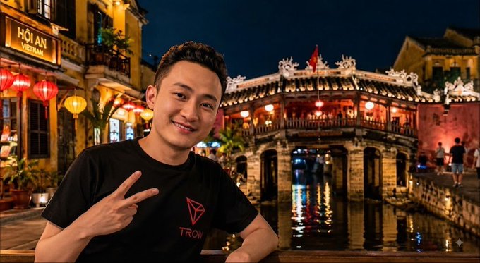 A photo of a smiling young man wearing a black TRON-branded t-shirt, making a peace sign in front of an illuminated Japanese covered bridge in Hoi An, Vietnam at night.