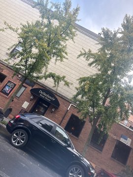 A street-level view of a brick building with a 'Dolce' awning over the entrance, a black SUV parked on the street, and a large green tree in front.
