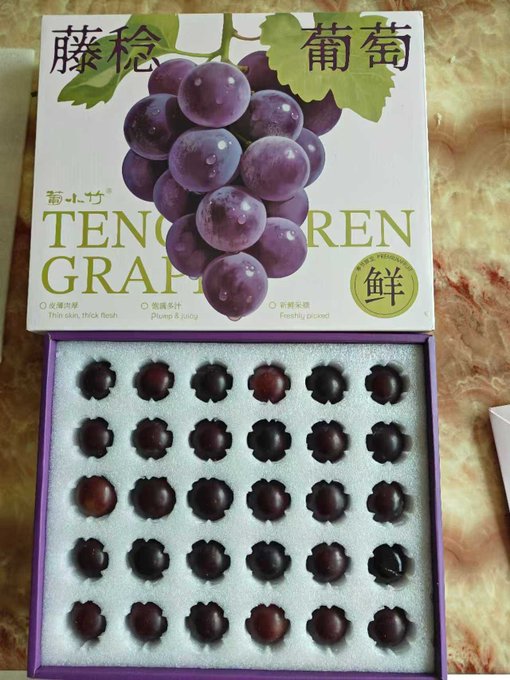 A box of 'Teng Ren' (藤稔) grapes with Chinese text describing them as 'Thin skin, thick flesh' and 'Plump & juicy,' showing purple grapes arranged in molded compartments in a white foam tray.
