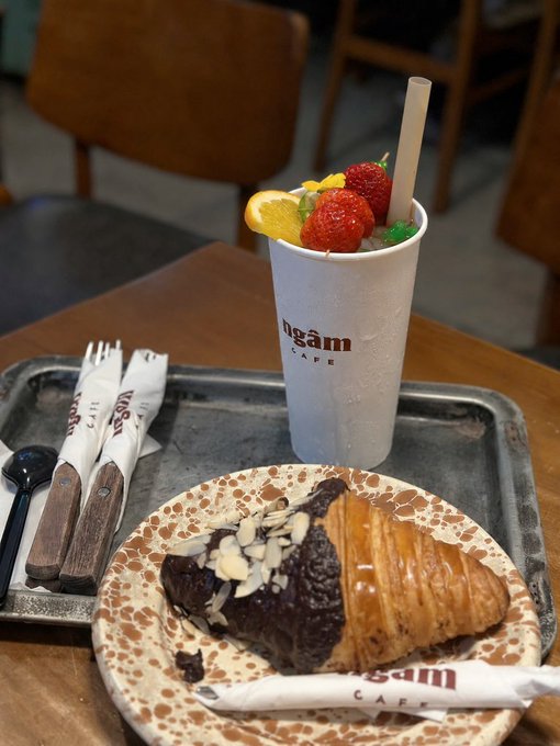 A café meal featuring a chocolate croissant topped with sliced almonds on a speckled plate, served alongside a cup garnished with strawberries and a lemon slice from 'ngẩm CAFÉ'.