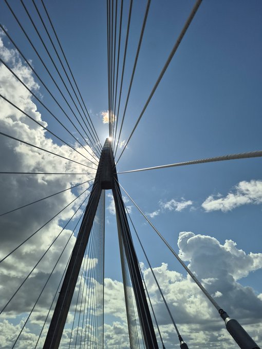 A low-angle photograph looking up at a cable-stayed bridge tower against a partly cloudy blue sky with sun flaring through the cables.