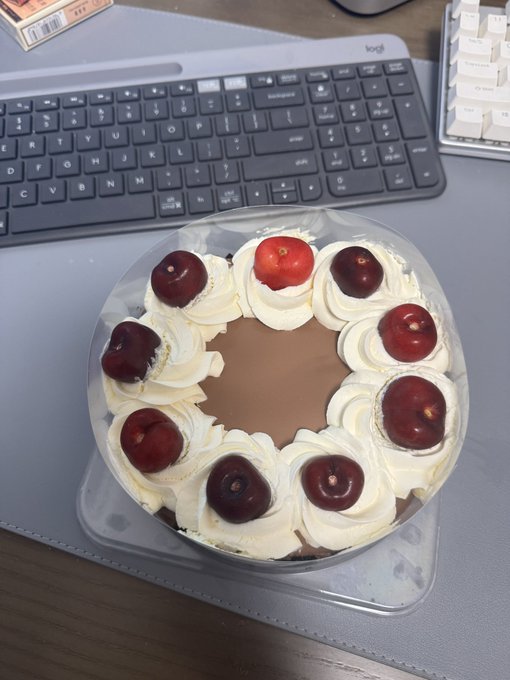 The image features a delicious chocolate cake with white frosting and cherries on top placed on a desk next to a computer keyboard. The cake appears to be a birthday cake, as indicated by the presence of a "Happy Birthday" message written on it. The cake is in a clear plastic container that sits on the desk. In the background, there are two keyboards - one near the bottom left corner and another towards the top right corner of the image.