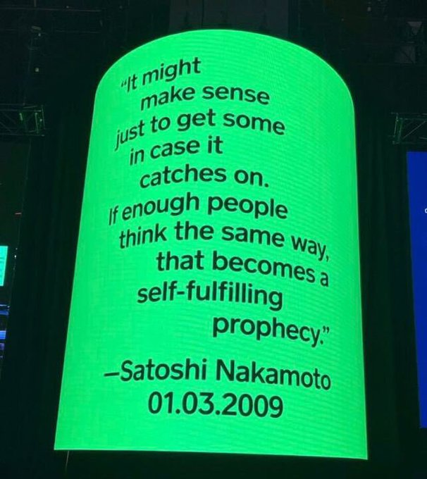 A glowing green digital display shows a quote attributed to Satoshi Nakamoto dated 01.03.2009: "It might make sense just to get some in case it catches on. If enough people think the same way, that becomes a self-fulfilling prophecy." The image directly echoes the post's reference to a self-fulfilling prophecy, visually highlighting the Bitcoin founder's words about collective belief shaping adoption.