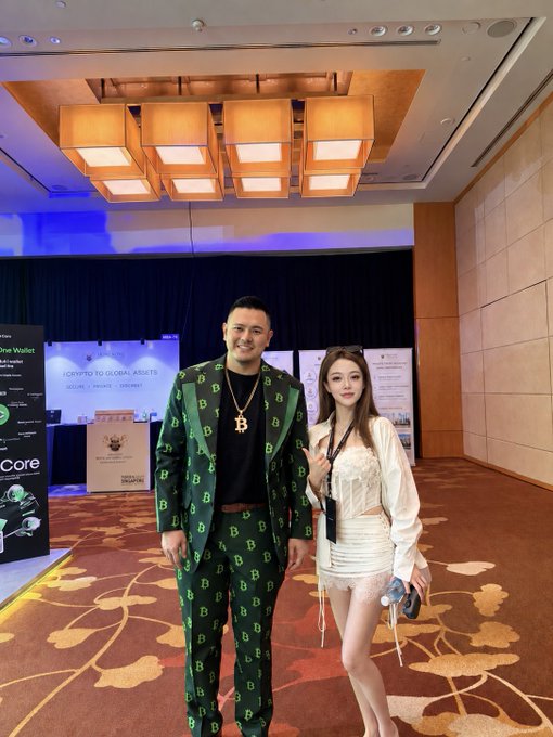 A man wearing a green Bitcoin-patterned suit and a Bitcoin necklace poses next to a woman in a white lace outfit at what appears to be a crypto conference, with a "Crypto to Global Assets" sign visible in the background. The post mentions this is at the Mixin Messenger booth (MB4-92, 4th floor), where attendees can claim giveaway surprises.