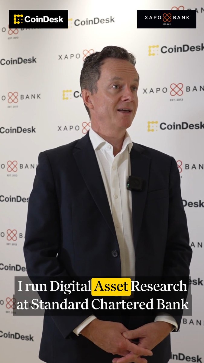 A man in a dark suit speaks at a CoinDesk event with Xapo Bank branding in the background, with on-screen text identifying him as running "Digital Asset Research at Standard Chartered Bank." The caption contextualizes this as Geoffrey Kendrick, Standard Chartered's head of digital asset research, sharing his bullish Bitcoin outlook including a $100K year-end target driven by returning ETF buyers and digital treasury narratives.