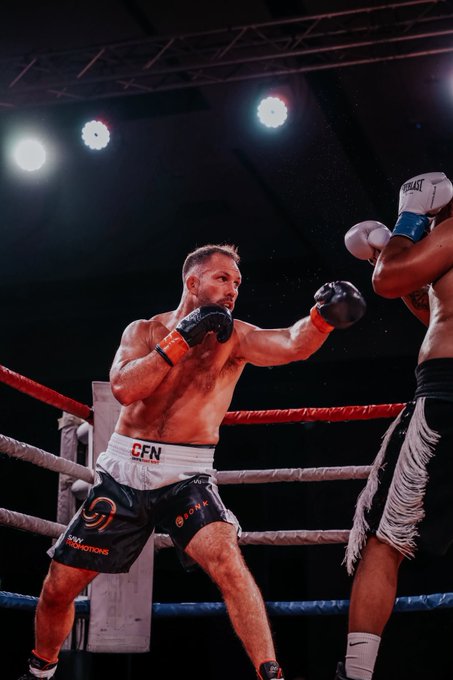 A focused boxer wearing "CFN" branded shorts throws a punch at his opponent during an intense match under bright arena lights. The image captures the raw energy and athleticism of combat sports, fitting for a dynamic sports-focused post.