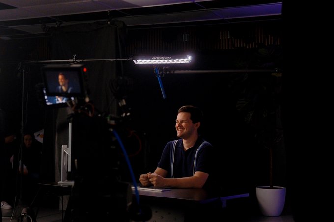 A man in suspenders sits at a desk smiling during a video shoot, with professional lighting and a camera monitor visible.