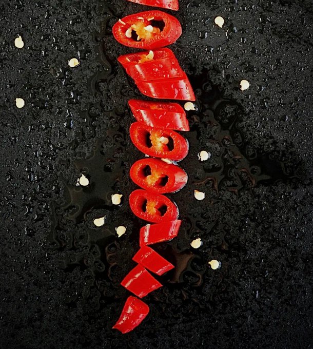 Sliced red chili pepper rings arranged vertically on a wet black textured surface, surrounded by scattered seeds.