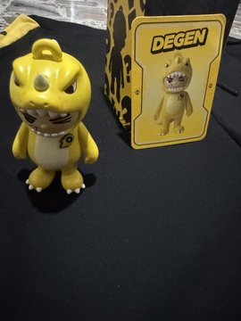 A small yellow monster-like figurine with a grumpy expression stands next to its packaging labeled 'DEGEN' on a dark surface.