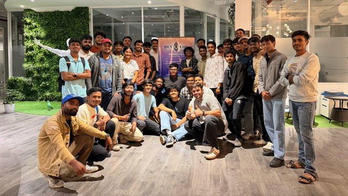 A group photo of approximately thirty young men posing indoors, with a 'DEV X CODERS' banner visible in the background.