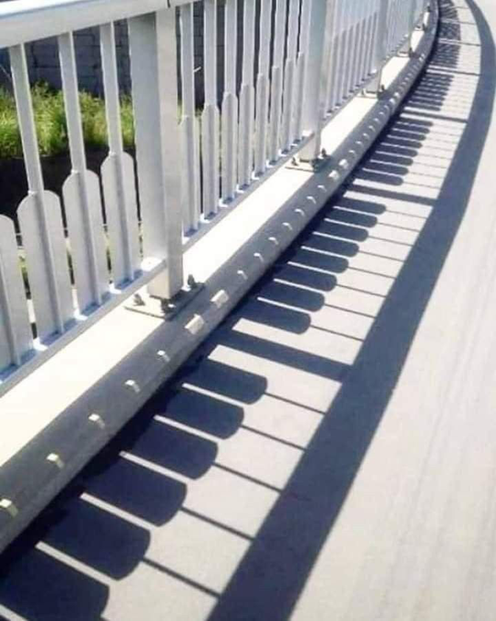 A photograph taken from a bridge showing the white metal railing casting a rhythmic shadow pattern on the pavement below, with a glimpse of greenery in the background.