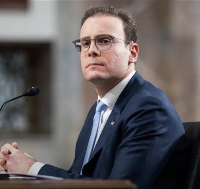 A man with glasses and a blue suit with an American flag pin sitting at a hearing or testimony with a microphone in front of him.