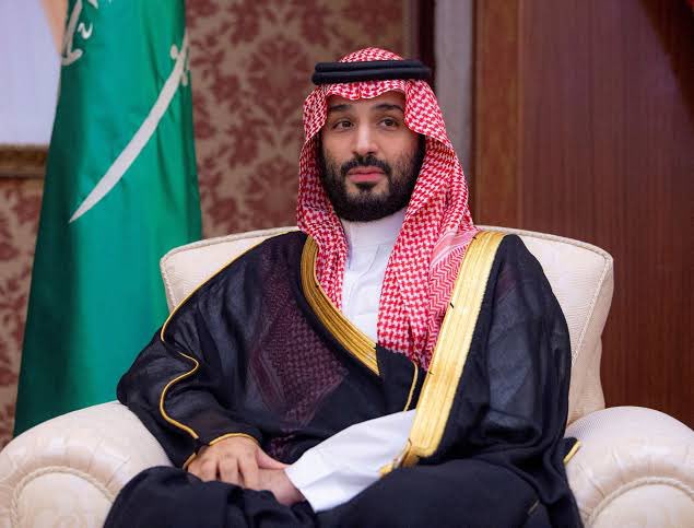 Saudi Crown Prince Mohammed bin Salman seated in traditional attire, wearing a red and white keffiyeh with a black bisht featuring gold trim.