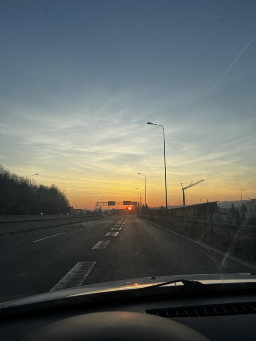 A view from inside a car driving on a highway at sunset, showing road lights, streetlamps, and a construction crane in the distance.