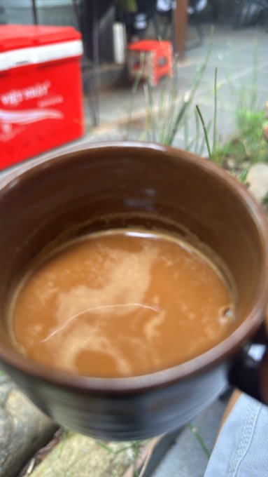 An outdoor close-up of a brown ceramic mug filled with milky coffee, with a red cooler and grass visible in the blurred background.