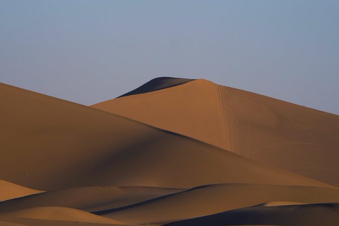 Smooth, sweeping sand dunes in a desert setting are illuminated by warm light, with subtle vehicle tracks visible on the steep dune face beneath a pale blue sky.
