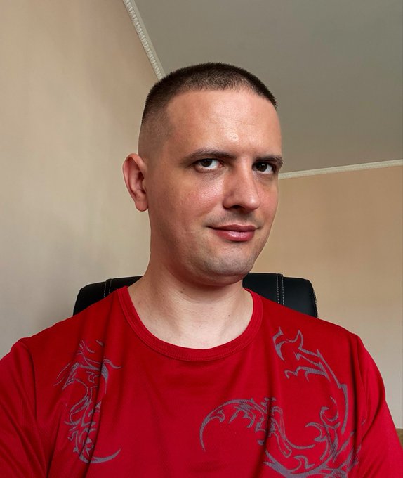 A man with a buzz cut wearing a red shirt with gray dragon designs sits in a chair against a beige wall.