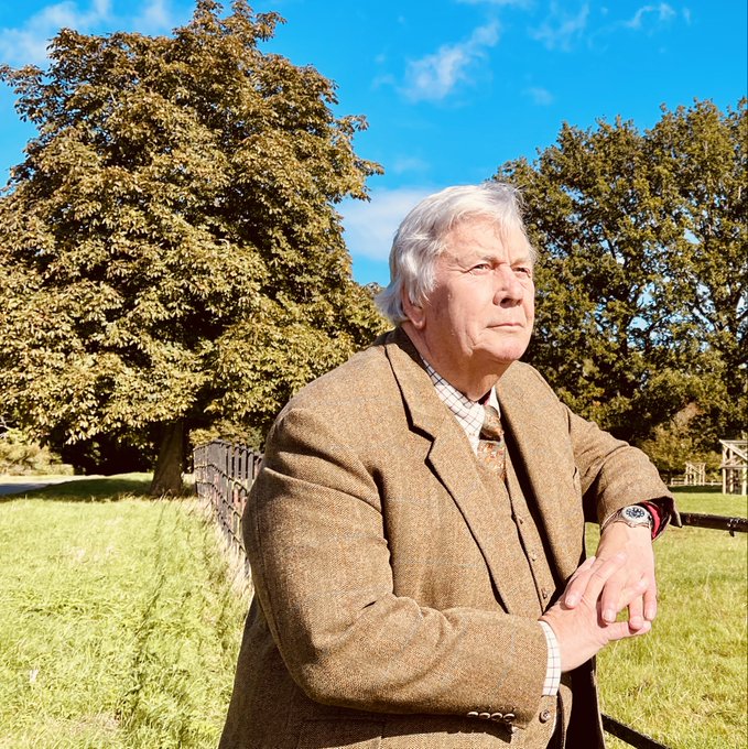 Outdoor portrait of an older gentleman with white hair wearing a brown tweed jacket, posed in front of large green trees in a grassy field.