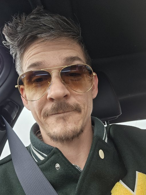 A man wearing aviator sunglasses and a dark varsity jacket smiles for a selfie inside a car. The image accompanies a post about loyalty and references "$ADA," though no text, numbers, or tickers are visible in the photo itself—the caption playfully suggests the man embodies the definition of loyalty.
