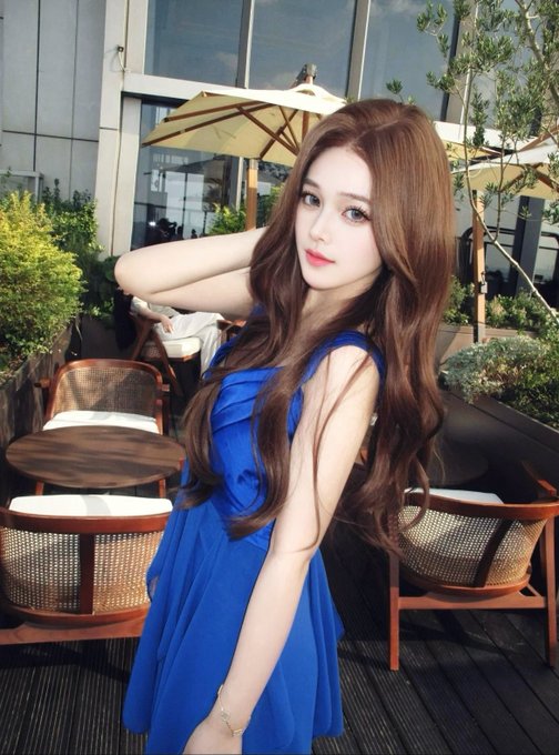 A young woman with long, wavy auburn hair poses gracefully at an outdoor café, wearing a vibrant blue dress, with patio furniture and umbrellas in the background—capturing a serene morning moment.