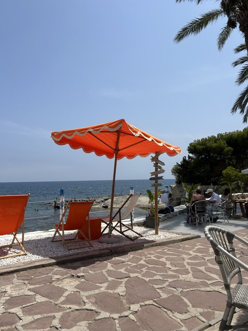 A sunny seaside promenade features an orange scalloped umbrella shading lounge chairs on a stone patio, with people seated at tables overlooking the calm blue sea.