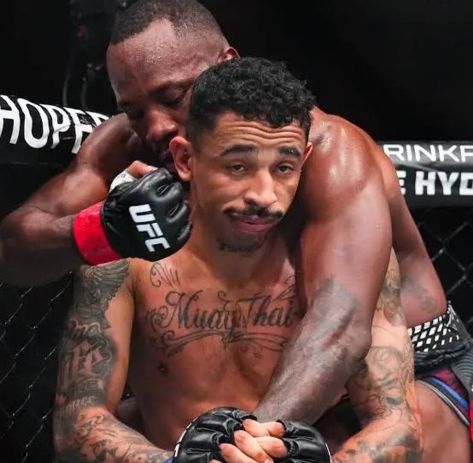 Two shirtless UFC fighters are grappling against the cage, with one fighter behind the other applying a hold, while the front fighter displays "Muay Thai" tattooed across his chest and wears black UFC gloves. The image humorously illustrates the post's caption of being "unbothered" by showing a fighter calmly tolerating a bear-hug rear choke attempt.