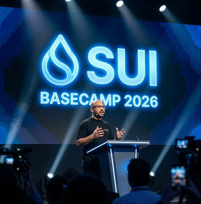 A man giving a presentation on stage at the 'SUI BASECAMP 2026' event, speaking at a podium with the blue illuminated SUI logo displayed on a large screen behind him.